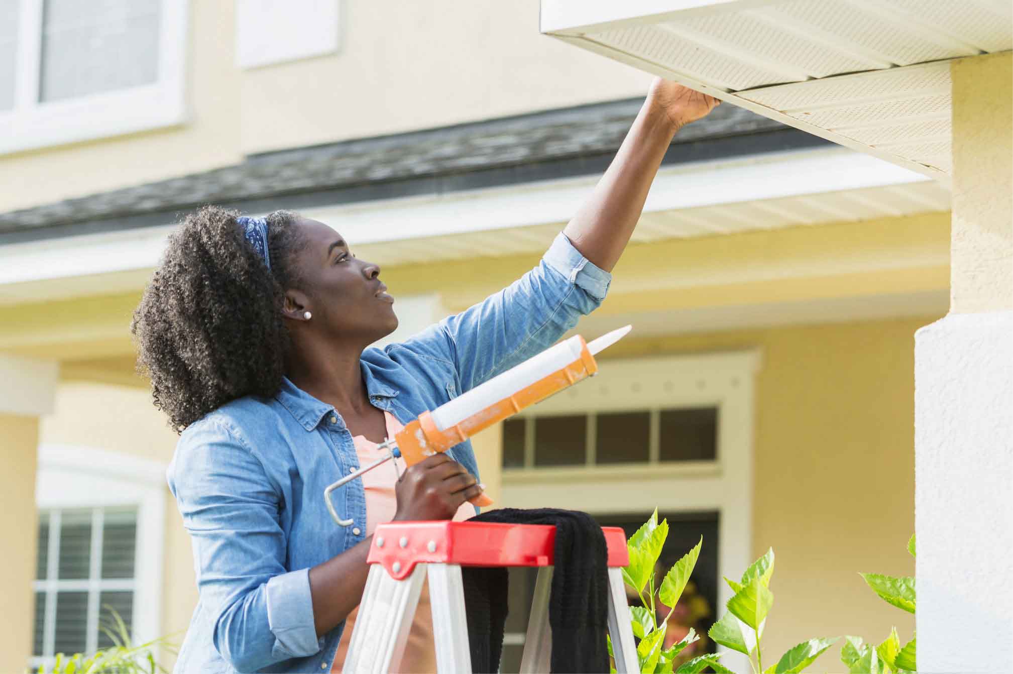 A woman in a denim shirt standing on a ladder while checking her gutter.
