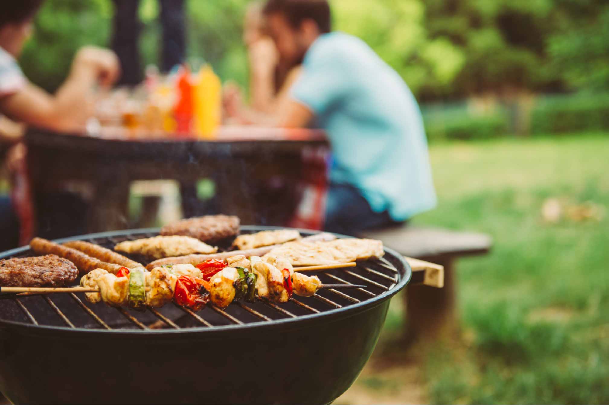 A close up of burgers and skewers on a barbecue with people sitting in the garden behind.