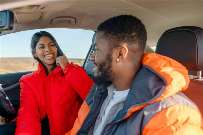 Young couple sat in front of a car. They are looking at each other and smiling.